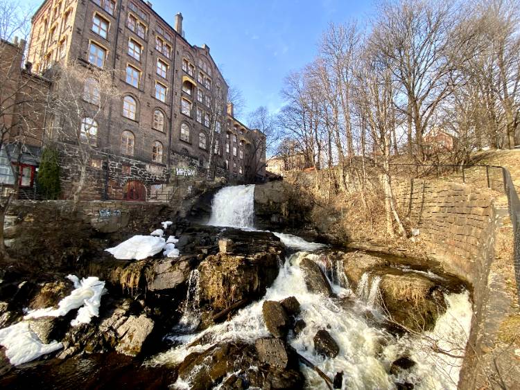 the akerselva river in oslo flowing downhill, with white water in several places. There is a large, brick building in the background.