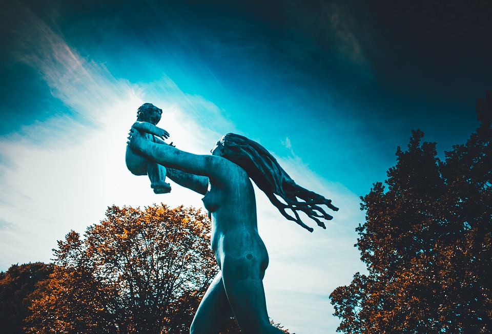 a statue of a womanwith long hair holding a baby up high, with trees with brown leaves in the background, and a blue sky above. 