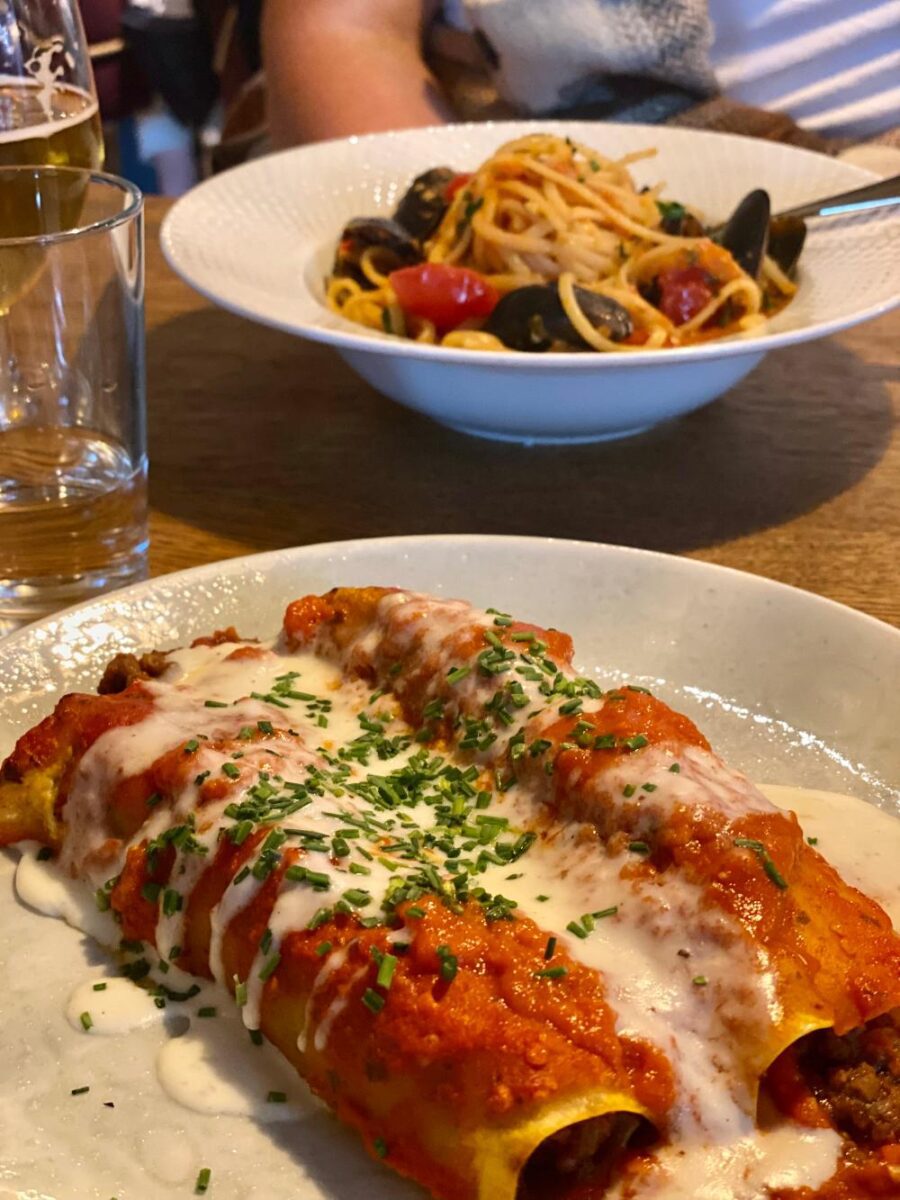 a plate of two pieces of cannelloni in the foreground, and a seafood spaghetti in the background, at a bergen restaurant with dark wooden tables