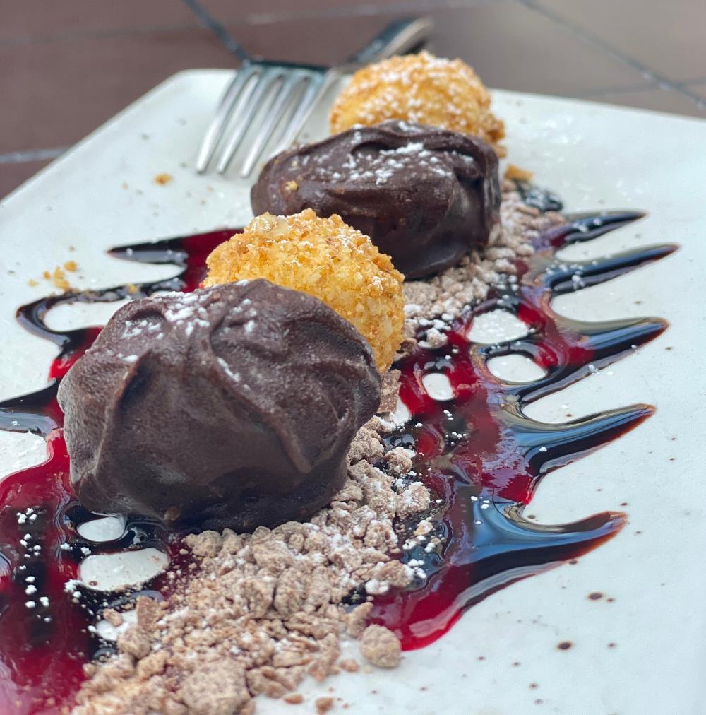 four cheesecake balls covered in hard chocolate on a white plate with a fork in the background leaning on the plate