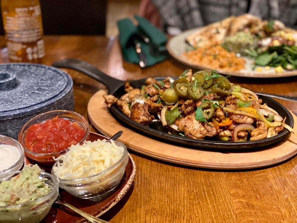 photo of a plate of chicken with vegetables and jalapenos, on a wooden table. In the foreground are small bowls of fajitas topping: cheese, salsa and guacamole.