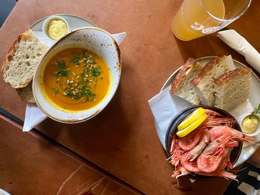 a flatlay photo of a bowl of orange soup with sourdough bread next to it, and a bowl of king prawns with bread next to it on a brown table.