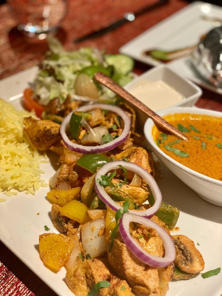 a chicken dish with a small bowl of orange sauce next to it. The dish consists of small pieces of chicken, cut vegetables and onion rings on top.