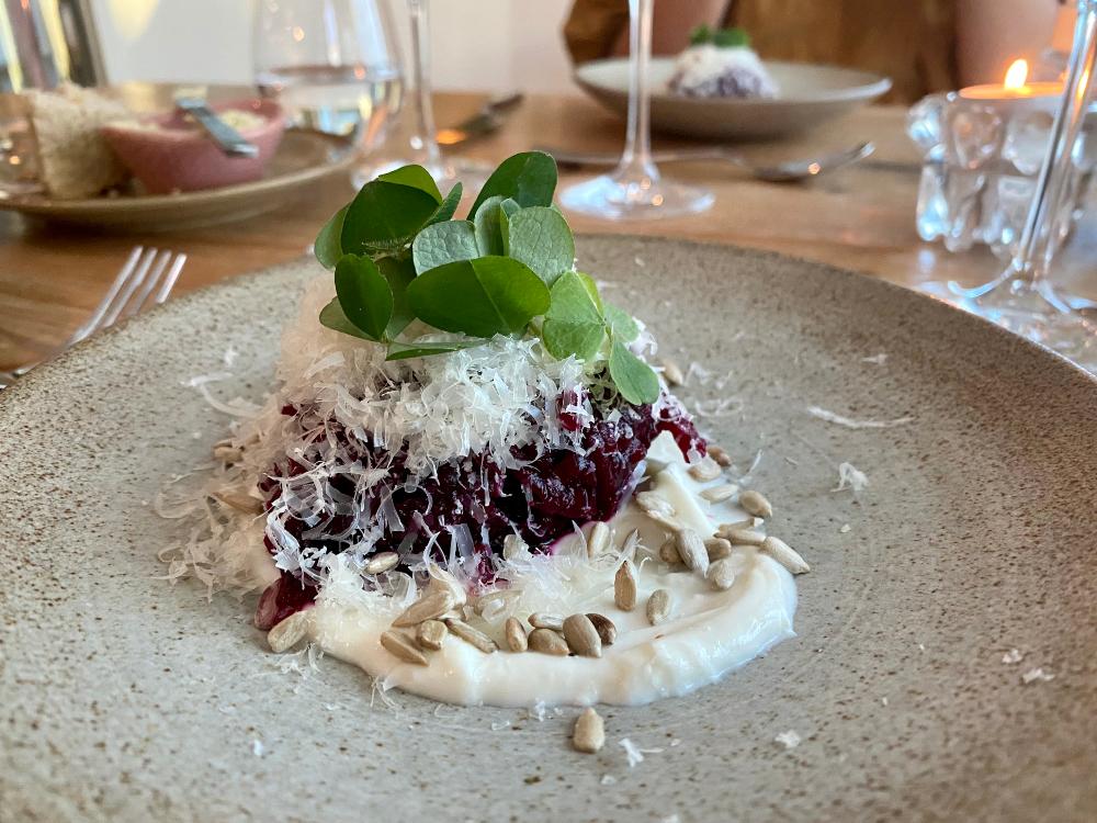 a dish at a fine dining restaurant, on a dark hand-made plate. the dish includes beetroot, grated goat's cheese and green garnishing. the restaurant is called Marg og Bein.
