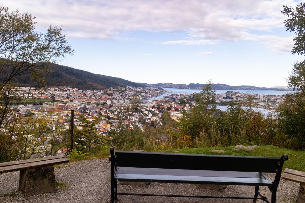 a green bench in the foreground overlooking a waterfront city with blue skies and clouds above. The city is bergen, norway