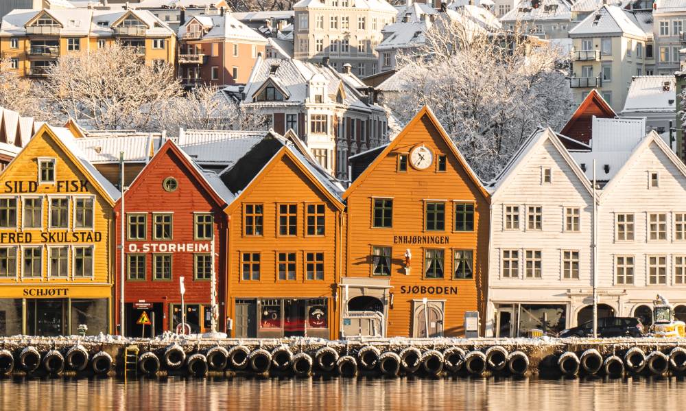 6 wooden houses lining the docks in Bergen, painted in yellow, red and white. The words Enhjørningen are painted in black on the middle, yellow house, indicating that this Bergen restaurant is inside.