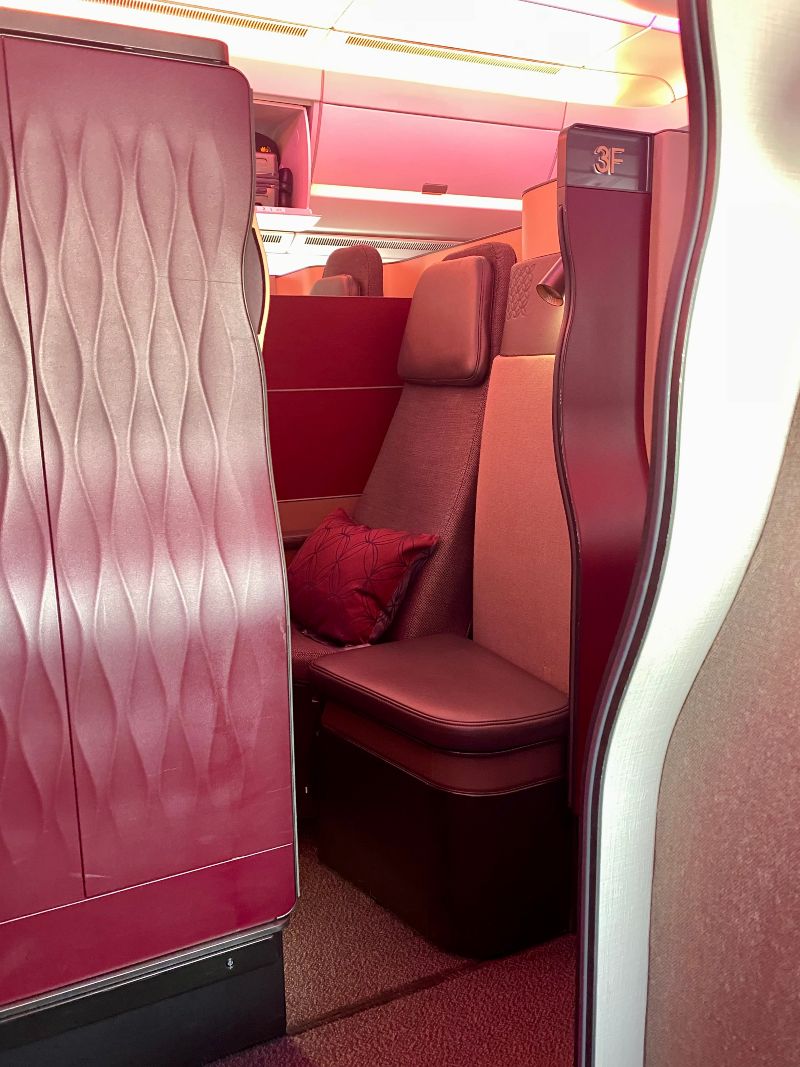 a sideways photo of the qatar qsuite, with the maroon door open. You can see a grey airplane seat through the open door, and a maroon pillow on the seat