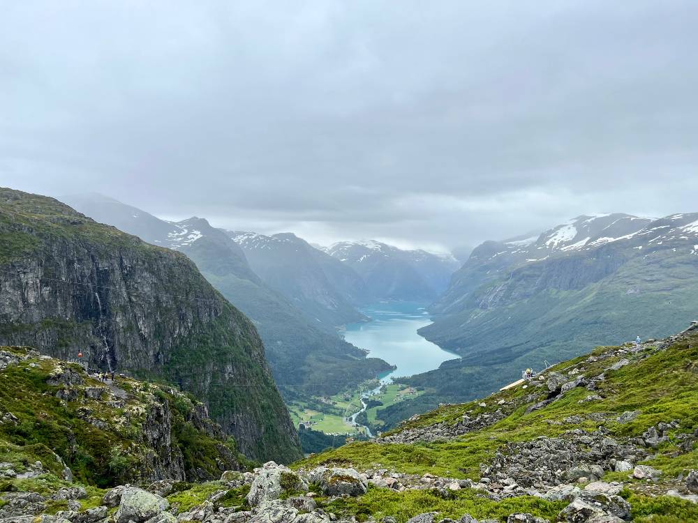 A view from a mountain top in Loen, Norway, showing a glacier lake and valley below, with high, snow-capped mountain tops surrounding the valley.