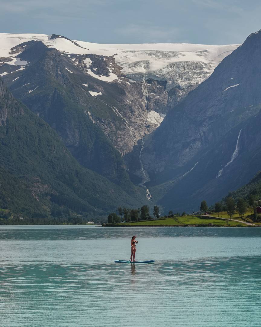 A woman on a SUP board on a bright green lake in Norway, with tall snow capped mountains in the far background.