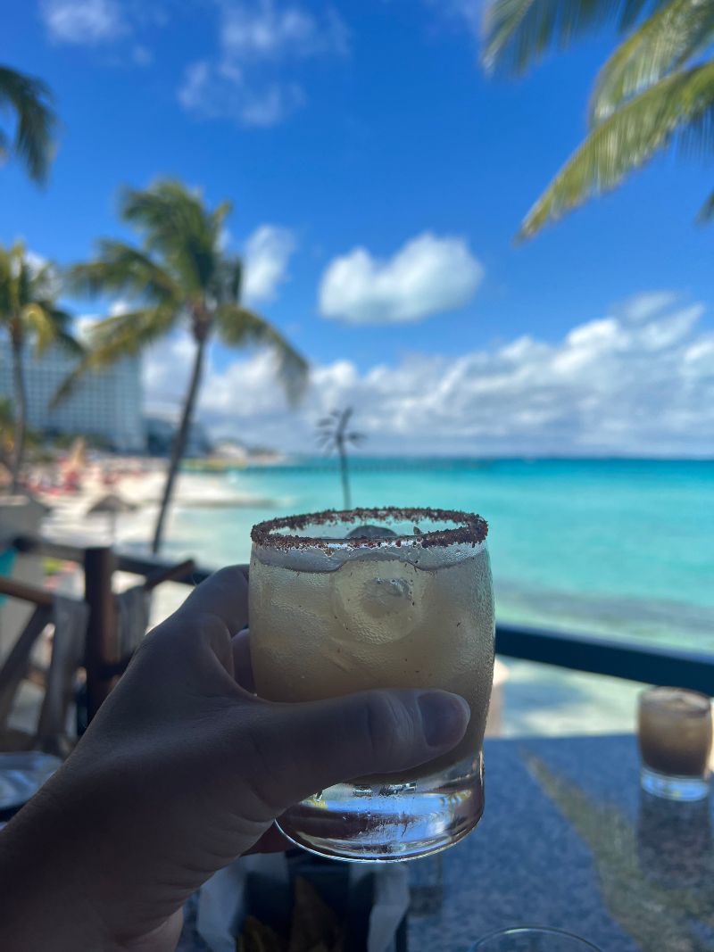 a white hand holding a yellowish, see through drink with a red salt rim. In the background is a bright blue ocean.