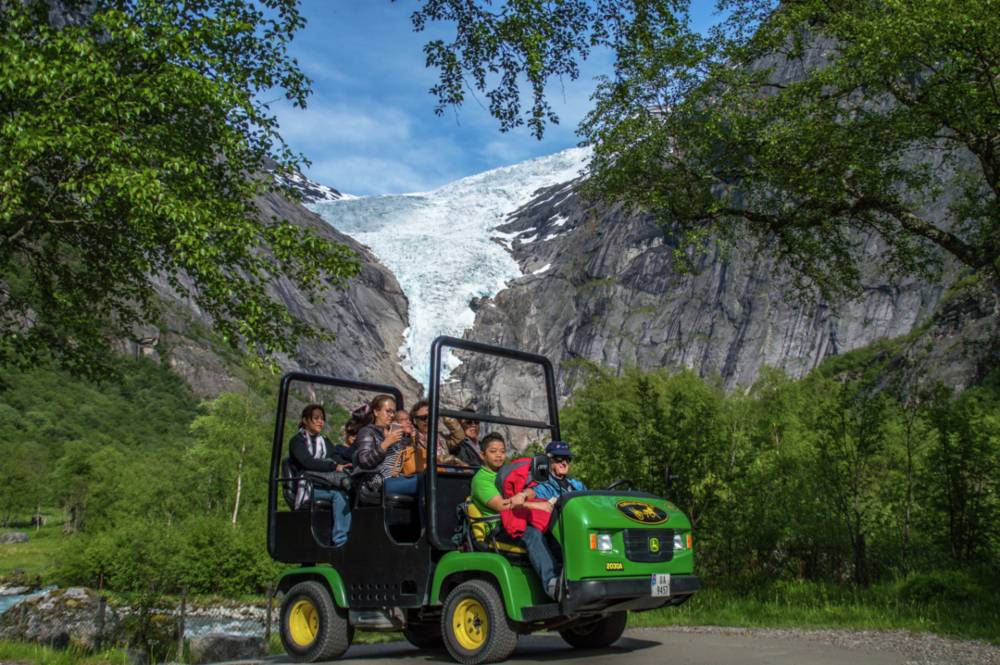 A bright green golf cart driving on a small road in the foreground, with greeneries and shrubs surrounding it, and a glacier in the background.