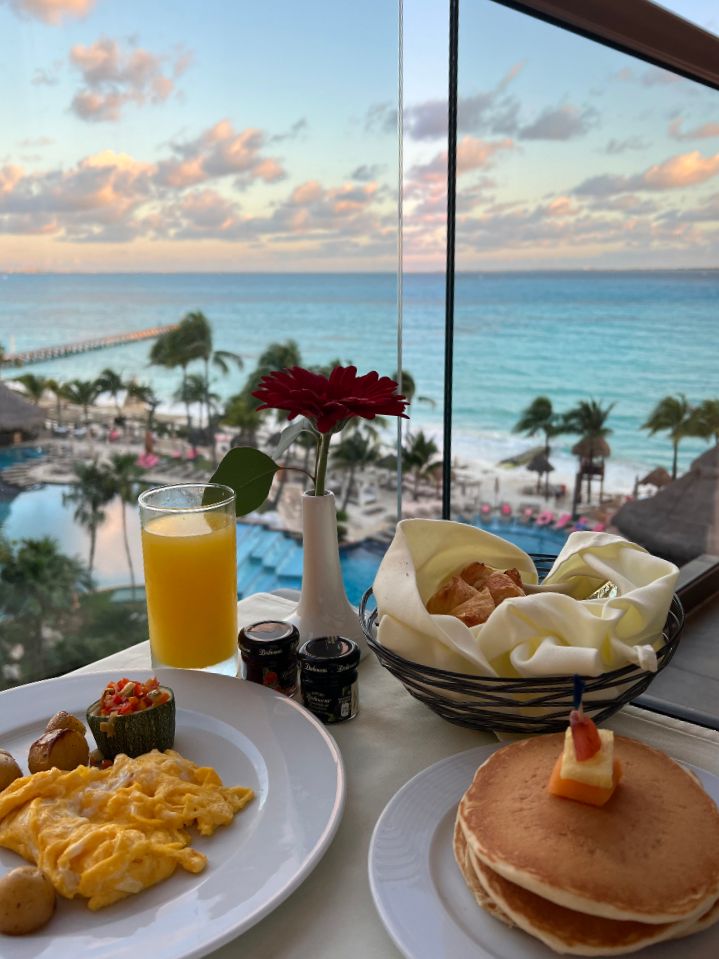 scrambled eggs, pancakes and orange juice on a small table in the foreground, with a blue ocean and pink skies in the background. This is a photo of a room service breakfast on a balcony in Cancun, Mexico.