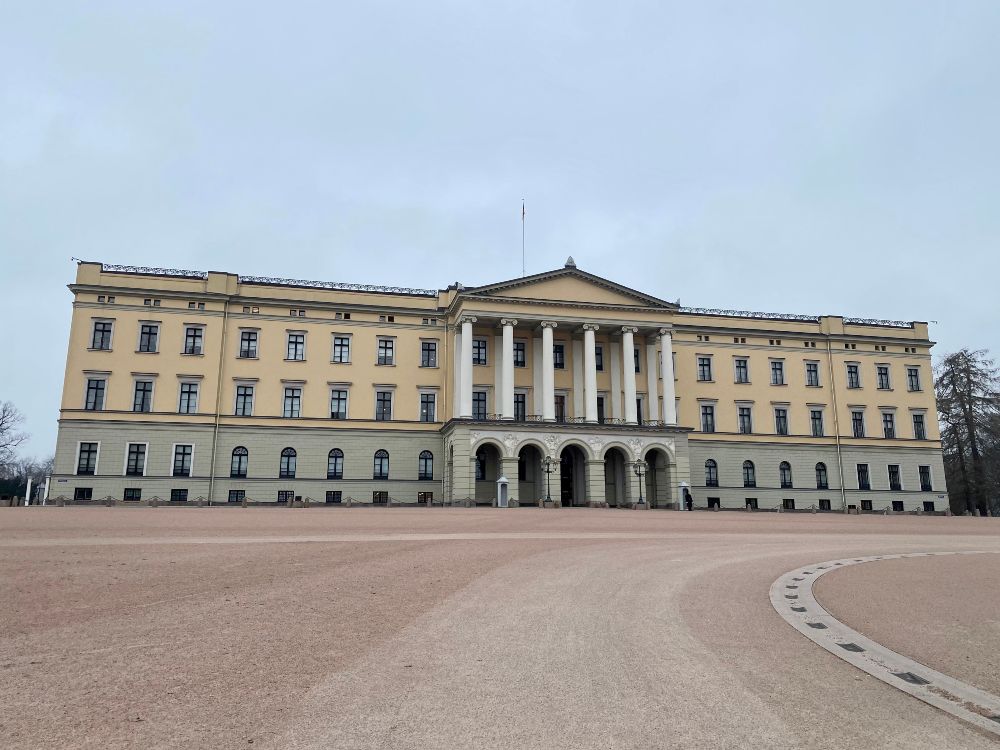 The Norwegian Royal Palace in Oslo, seen from the front. This is a large, light yellow building, with 6 massive pillars above the main entrance in the middle.