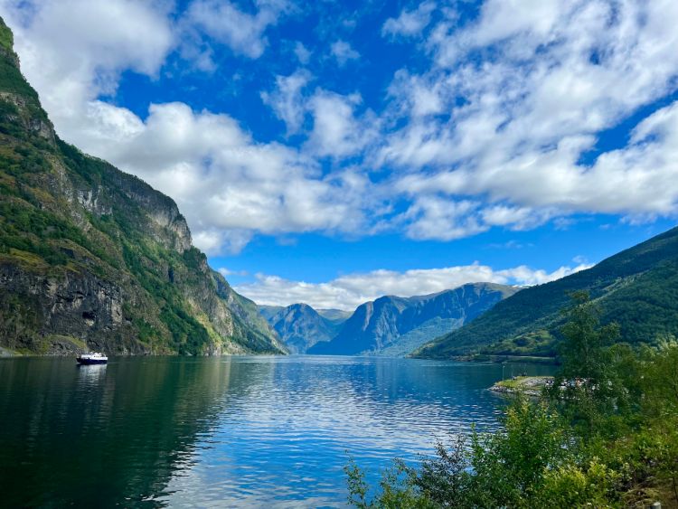 A beautiful photo of a quiet fjord in Norway, with tall mountains on either side and blue, cloudy skies above. There is a small boat sailing across the fjord to the left of the photo. This area is part of the Norway in a nutshell tour.