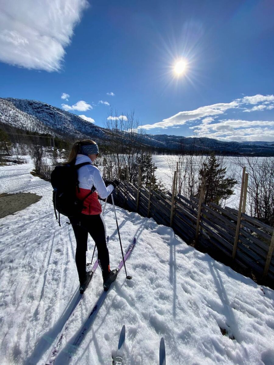 A girl on skis with a white and red jacket, and a black backpack on her pack. She is looking over a frozen, snowcovered lake, and the sun is shining bright above it.
