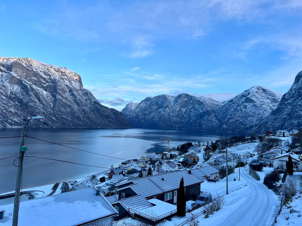 A winter fjord landscape in western Norway. Snow-covered roofs lining a fjord, with steep, snow-capped mountains on the other side.