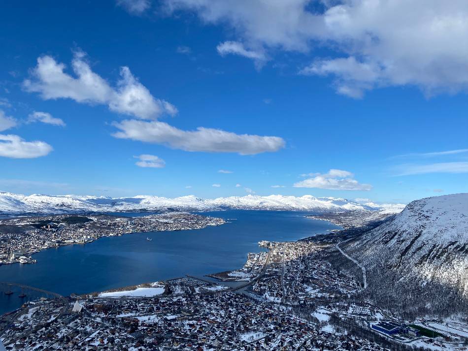 the view of the city of tromsø from the fjellheisen cable car, with blue skies above and snow on the mountains and in the city