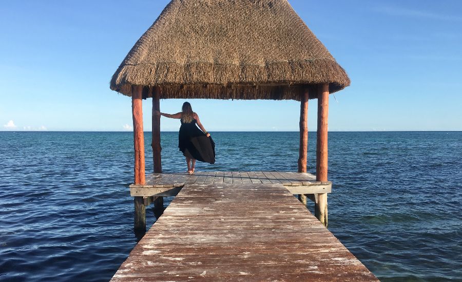 A girl in a black dress standing on a pier in Tulum, Riviera Maya, looking over the dark blue ocean.