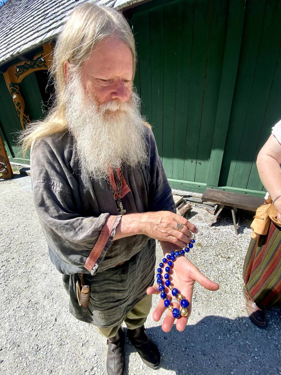 A Viking chieftain with a long white beard holding up a blue, beaded necklace to show the camera. He is wearing Thor's hammer around his neck, a famous nordic symbol.