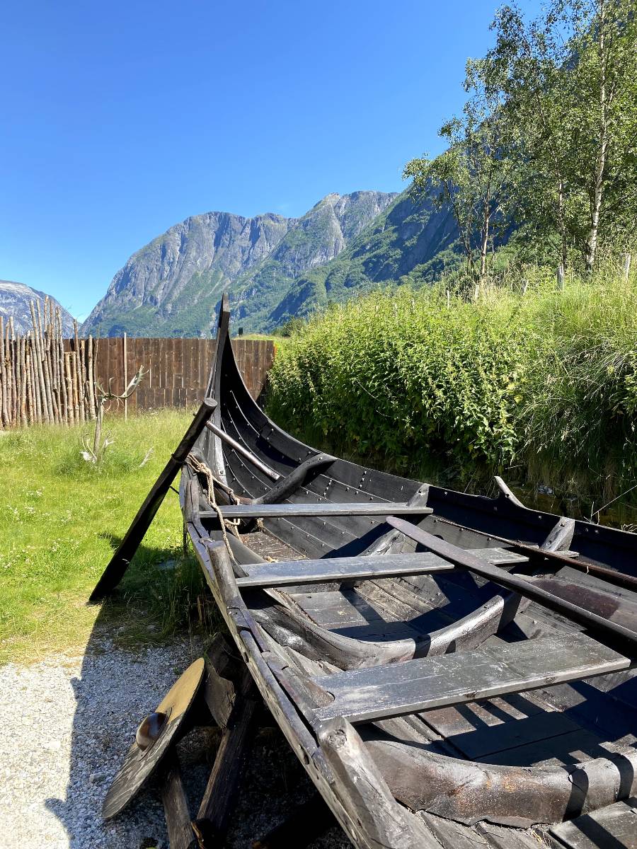 A photo of a replica of a small Viking longship, with a dark wooden ship taking up the majority of the image. In the background is green grass and blue skies.