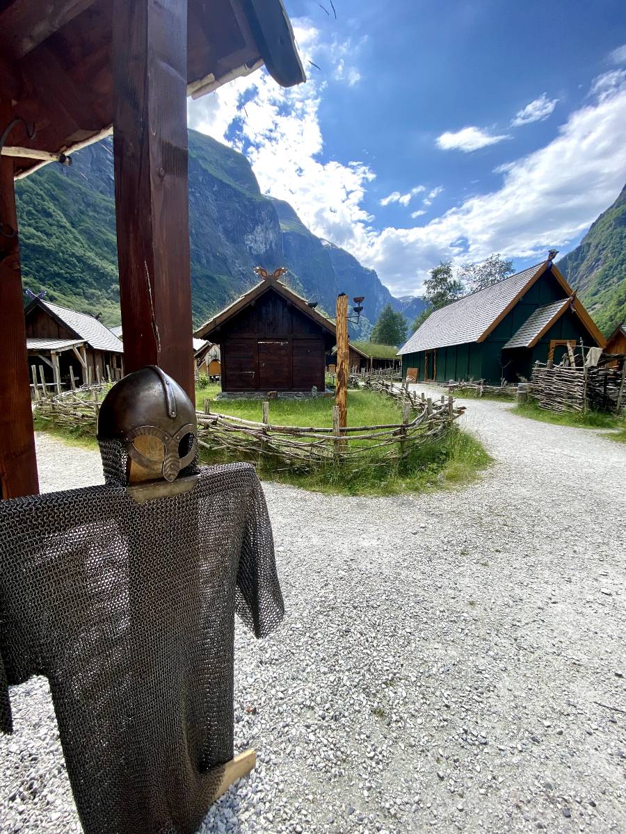 A viking helmet and armor hanging on a wooden mannequin, with a wooden fence, garden and viking houses in the background. The helmet (without horns) is an important Viking symbol.