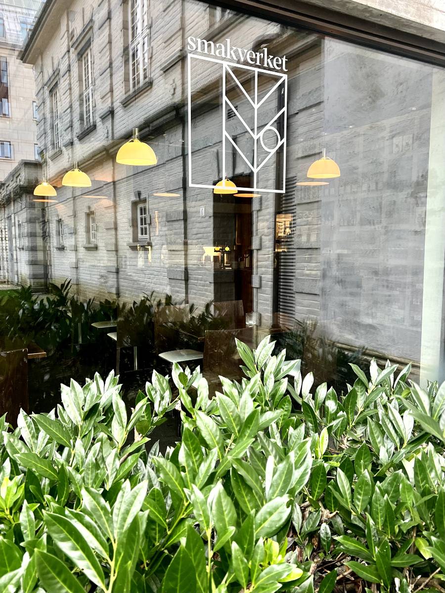 A glass window with a logo saying Smakverket on it. At the bottom of the photo are some bushes, in front of the cafe window. You can see some ceiling lamps and cafe chairs and tables through the window.