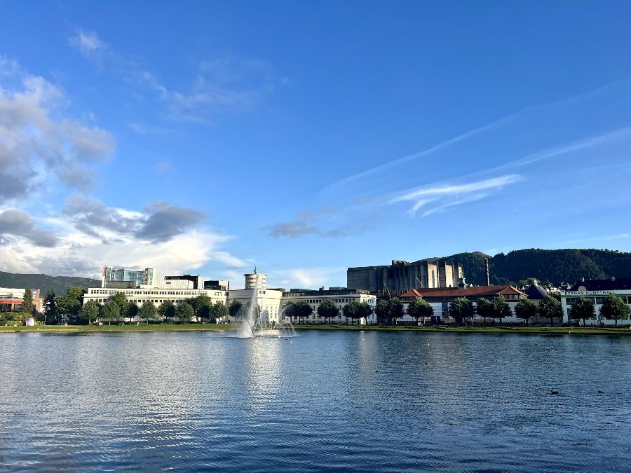 The city of Bergen seen across a small man-made lake with a fountain spurting water in the middle. The sky is blue, with some white clouds above.