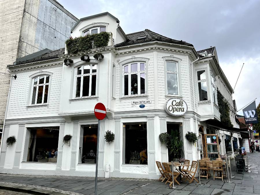 A white wooden building with some small tables and chairs outside, and a sign saying Cafe Opera above the entrance.