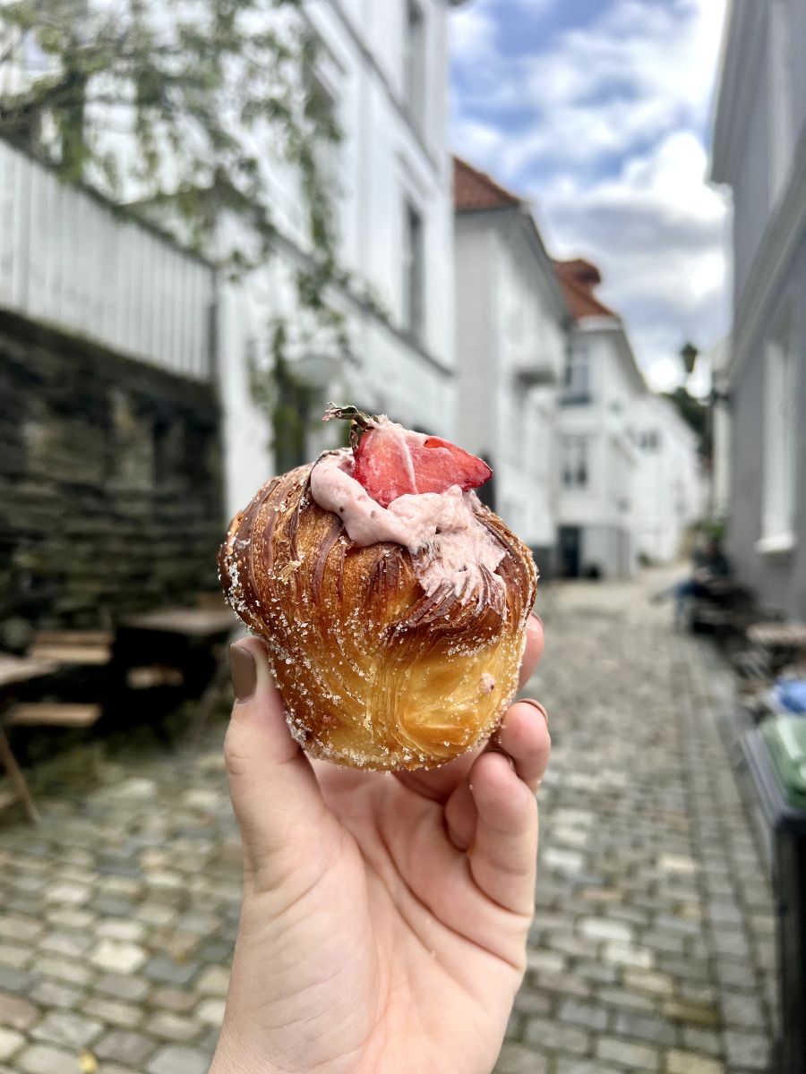 a hand holding a cruffin with a strawberry on top in front of a small cobbled street in Bergen, Norway. The street is blurred and the cruffin is in focus.