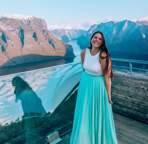 A girl in a white top and blue long skirt smiling at the camera with her arms down along her sides, on a viewpoint overlooking a Norwegian fjord. This is Lisa Stentvedt, author and travel blogger from Norway