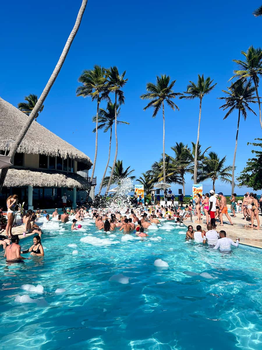 A photo of a hotel pool filled with people swimming and dancing, and foam being sprayed out of a cannon by the pool. Foam is also floating in the pool, and there are palm trees in the background.