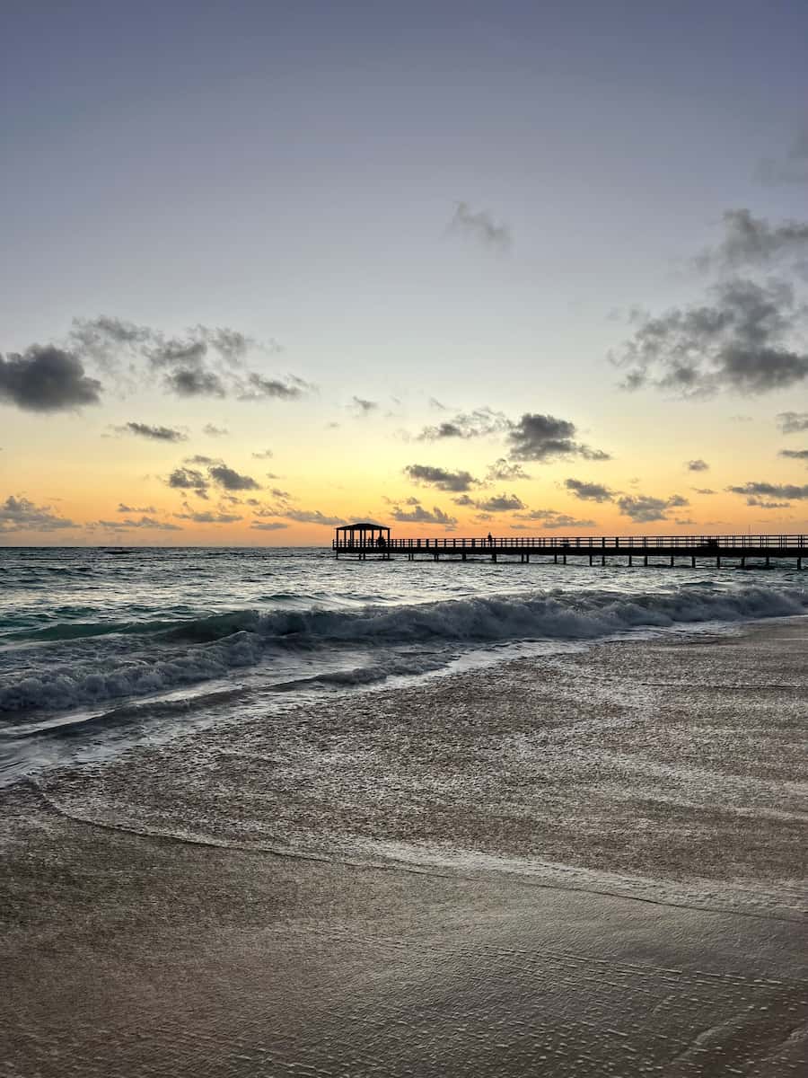 A photo of a wooden pier jutting into the ocean, with orange skies in the background and waves in the foreground.