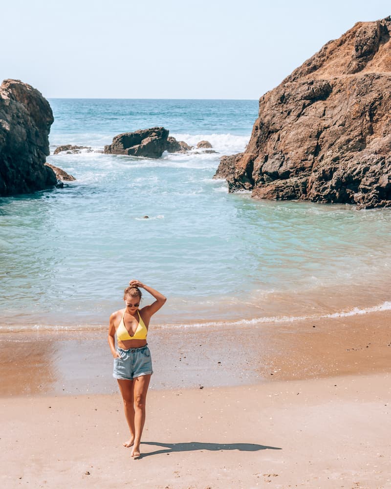 A woman in a yellow bikini top and jeans shorts standing on a small beach in New zealand, looking down at the sand. There are some rocks in the water in the background, and waves crashing over the rocks.